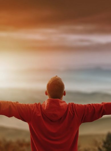 Man with arms spread looking at mountains vivid photo with layered mountains valley in front. Man has his back facing the camera with mountains in background and sun flare effect